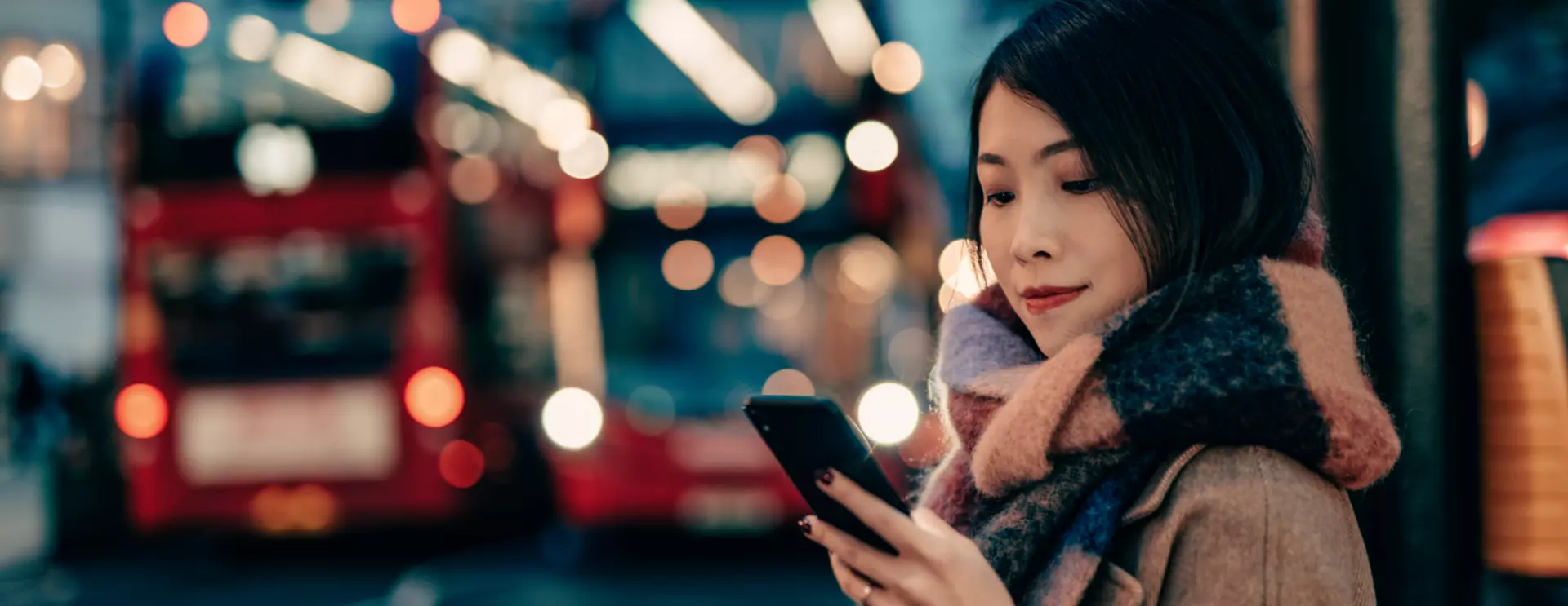 Woman wearing a wool coat and scarf looks at her phone with two red double-decker buses in the background in London, England 